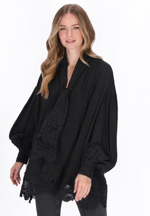 Femme aux cheveux longs et ondulés portant un chemisier noir ample avec des détails en dentelle sur les manches et l'ourlet, debout devant un fond blanc.