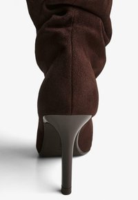 Hnedé semišové vysoké podpätky s špicatou špičkou a štíhlym, elegantným podpätkom. Tkanina má jemnú textúru a je zdobená zhlukovaním na lýtku.