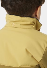 Chaqueta amarilla con cuello alto, de textura suave, detalles de botones a presión y ribete oscuro en los bordes.