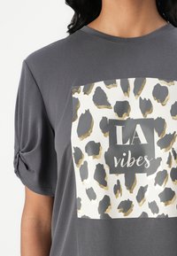 T-shirt gris à manches courtes avec un imprimé en avant de taches de léopard en or et gris, accompagné du texte "LA vibes" dans un encadré noir. Détail de manche nouée.