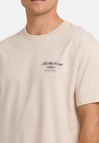 Beige T-shirt met korte mouwen van katoen, met een ronde halslijn en een kleine grafische print op de linkerkant van de borst met tekst en designelementen.