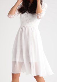 Vit klänning med en spetslivstycke och trekvartsärmar. En flödande chiffongkjol faller nedanför knäet och skapar en lager-effekt.
