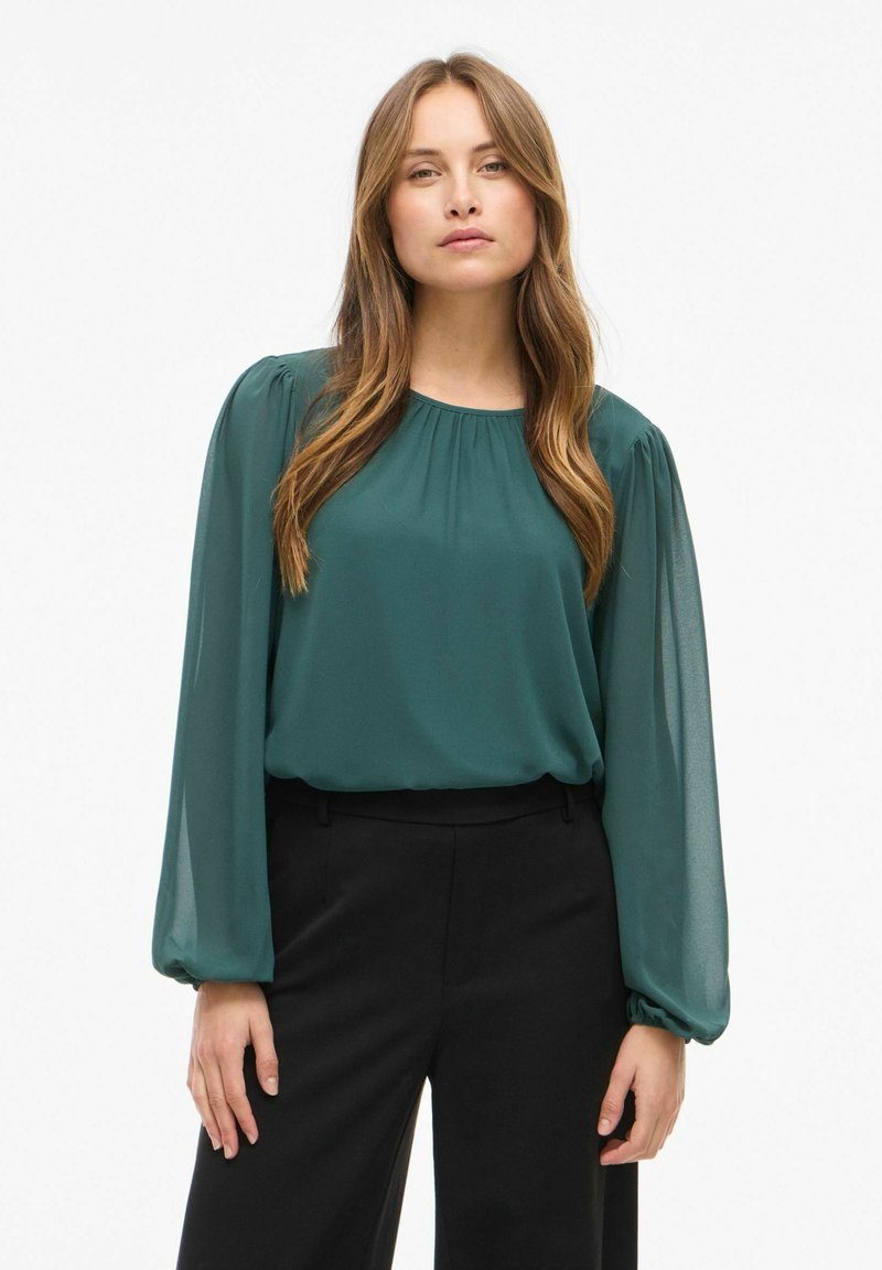 Blusa color turchese con maniche lunghe e arricciate, scollatura rotonda e vestibilità morbida. Abbinata a pantaloni neri, con una texture liscia e un design sartoriale.