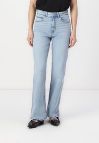 Light blue denim flared jeans with a high waist, five pockets, faded areas, and subtle whiskering details. Paired with black pointed shoes.