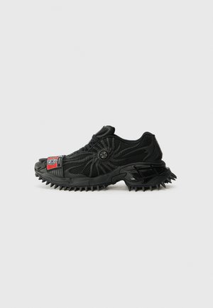 Black athletic shoe with a textured mesh upper, rubber outsole featuring pronounced spikes, and a distinctive red and black logo patch.