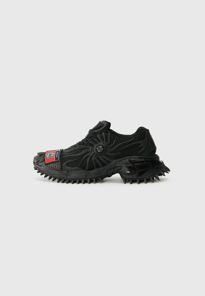 Pantofila sport neagră cu un superior din plasă texturată, talpă din cauciuc cu cuie pronunțate și un patch logo distinctiv roșu și negru.