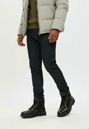 Uomo che indossa una giacca a piumino grigio chiaro, maglione verde oliva, jeans skinny scuri e stivali da combattimento neri con lacci, in piedi su uno sfondo bianco.