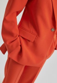 Oransje blazer og bukser med en glatt tekstur. Blazeren har en enkelt knapplukking og en sidelomme, skreddersydd design.