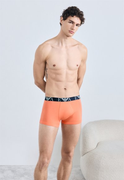 Orange boxer briefs with a navy elastic waistband displaying the brand logo. The fabric appears smooth and fitted, enhancing the shape.
