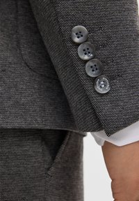 Manica di blazer grigio con motivo, completa di quattro bottoni scuri, realizzata in un tessuto testurizzato e con una tasca esterna. Polsino bianco visibile sotto.