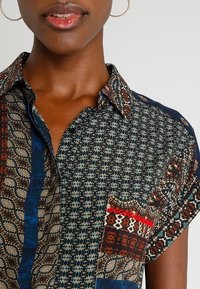 Chemise à manches courtes présentant des motifs variés dans des tons foncés, des designs élaborés, et un col boutonné avec des textures contrastantes.