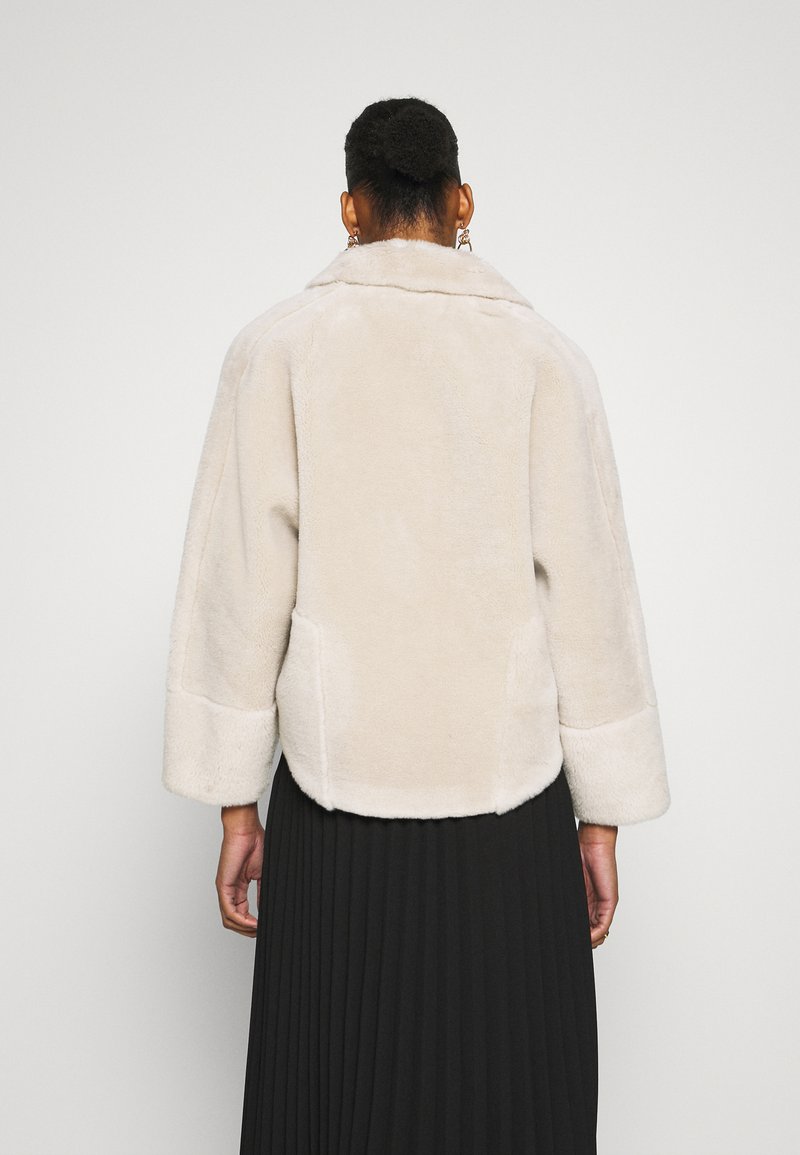 Veste en fausse fourrure beige clair avec des manches larges, un col et un ourlet arrondi. Texture lisse et design minimaliste.