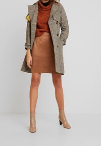 Trench-coat à motif pied-de-poule avec doublure jaune, pull en tricot marron, jupe en daim marron et bottines beiges à talon bloc.