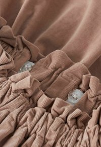 Gros plan sur une ceinture élastique beige ajustable avec des boutons transparents et un détail de tissu froncé sur un vêtement en velours côtelé.