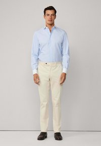 Uomo che indossa una camicia da uomo a maniche lunghe azzurro chiaro, pantaloni beige e mocassini marrone scuro, in piedi contro uno sfondo semplice.