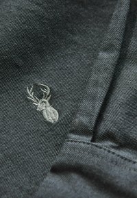 Tummanharmaa kangas, jossa on yksityiskohtainen brodeeraus pienestä peuran päästä, jossa on sarvet, sauman lähettyvillä näkyvillä ompeleilla.