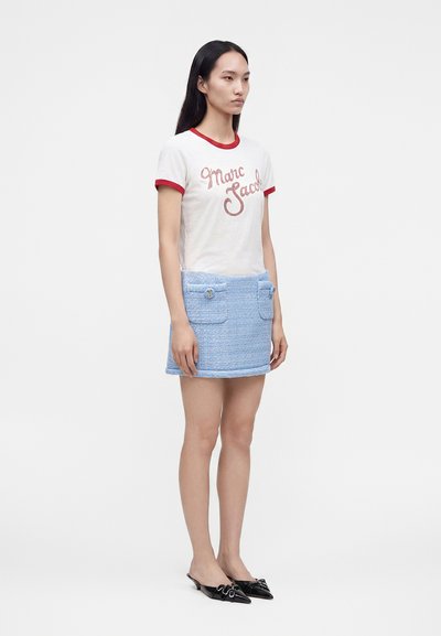 Γυναίκα που φοράει λευκό τ-shirt με κόκκινη λεπτομέρεια και κείμενο "Marc Jacobs", ανοιχτό μπλε μίνι φούστα με τσέπες και μαύρα μυτερά παπούτσια με λουράκι.