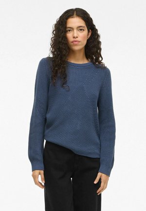 Femme aux longs cheveux bouclés portant un pull en maille texturée bleu et un pantalon noir, se tenant devant un fond blanc uniforme.