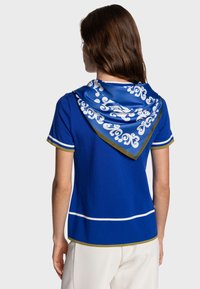 Haut bleu à manches courtes avec des accents blancs et dorés, accompagné d'une écharpe bleue à motif drapée dans le dos. Texture lisse, coupe ajustée.