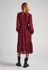 Žena v dlouhém burgundském midi šatech s béžovým diamantovým vzorem, v černých botách do půli lýtek, stojící na jednobarevném pozadí.