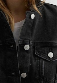 Svart denimjacka med silverfärgade tryckknappar, klassisk krage och subtila sömdetaljer. Tygets struktur är slät med en lätt sliten look.