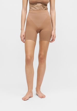 Vrouw draagt hooggetailleerde nude shaping shorts die tot halverwege de dij reiken, staat blootsvoets tegen een effen lichte achtergrond.