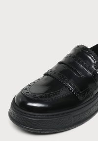 Μαύρο γυαλιστερό δερμάτινο παπούτσι brogue με διακοσμητικές διάτρητες τρύπες και παχιά υφή στην σόλα, εμφανιζόμενο σε κοντινή λήψη με λεπτομέρειες από μπροστά και πλευρά.