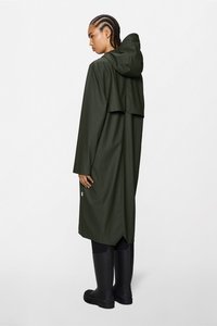 Olivengrøn regnfrakke med lang silhuet, der har hætte og delt rygdesign. Båret med sorte gummistøvler.