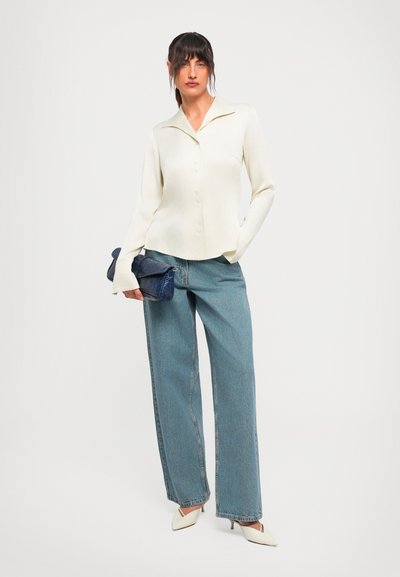 Woman wearing a cream button-up blouse, loose blue jeans, white heels, and holding a navy blue clutch against a plain white background.