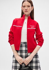 Chaqueta roja con cremallera y el logo "tommy girl", con detalles blancos y puños acanalados. Combinada con una falda de cuadros en blanco y negro y un bolso.