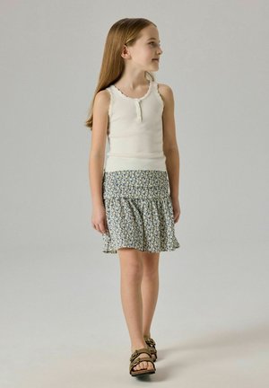 Nuori tyttö, jolla on pitkät hiukset, valkoinen hihaton paita, kukkakuvioinen hame ja ruskeat sandaalit, seisoo ja katsoo sivulle.