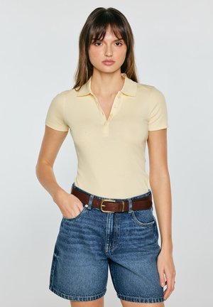 Młoda kobieta z prostymi włosami, ubrana w żółtą koszulkę polo z krótkim rękawem, jeansowe szorty i brązowy pasek, stoi z jedną ręką w kieszeni.