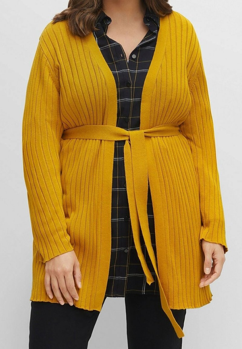 Musztardowożółty prążkowany kardigan z długimi rękawami i paskiem do wiązania, noszony nałożony na czarną i niebieską koszulę w kratę. Widoczna jest teksturowana tkanina.