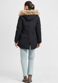 Doudoune noire avec une capuche en fausse fourrure, présentant une fente sur le côté et un design structuré. Associée à un jean délavé bleu clair et des bottes texturées.
