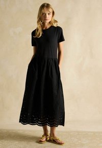 Melnas kleitas ar pieguļošu augšu un plūstošu svārku, kas rotāta ar acu izgriezumiem apakšmalā. Modelis valkā dzeltenas flip-flops. Mīksta, viegla auduma.