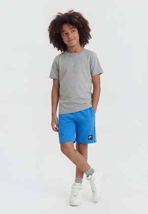 Παιδί με σγουρά μαλλιά ντυμένο με γκρι μπλουζάκι, μπλε σορτς Nike, γκρι κάλτσες και λευκά αθλητικά παπούτσια, στέκεται με σταυρωμένο το ένα πόδι.