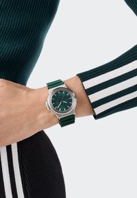 Zöld szilikon szíjas óra ezüst fém tokkal, sötétzöld számlappal és ezüst órjelekkel. Egy bordázott pulóveren viselve.