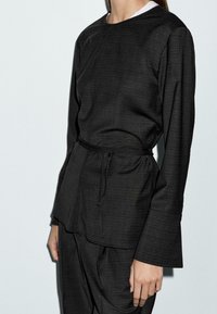 Mörkgrå texturerad topp med rund halsringning, långa ärmar och en knytning i midjan. Utsvängda ärmavslut och minimalistisk design.