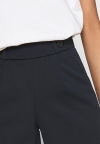 Tmavě modré kalhoty s hladkým povrchem, s ukrytou přední knoflíkovou západkou a bočními sklady. V kombinaci s bílým tričkem.