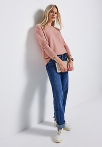 Jasnoróżowy sweter z prążkowanymi mankietami, zestawiony z luźnymi niebieskimi dżinsami i białymi sneakersami. Trzyma w plecionej kopertówce. Minimalistyczne tło.