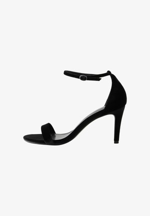 Sandalo con tacco alto in suede nero, con una sottile cinghia alla caviglia, punta aperta e una silhouette sinuosa e liscia. Presenta una chiusura a bottone sulla cinghia.