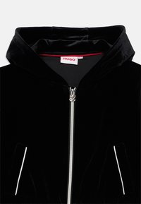 Giacca con cappuccio in velluto nero con zip argentata, caratterizzata da piping bianco sulle tasche. L'etichetta riporta la scritta "HUGO" in rosso. Texture morbida.