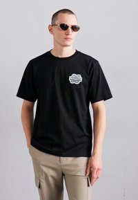 T-shirt en coton noir à manches courtes et col rond, avec un design graphique sur la poitrine. Porté avec un pantalon cargo beige.