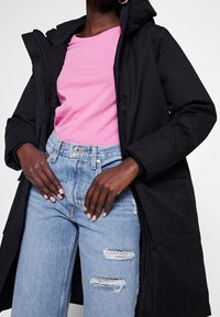 Chaqueta negra con capucha sobre una camiseta rosa, combinada con vaqueros claros rasgados. La chaqueta tiene una cremallera y bolsillos frontales.