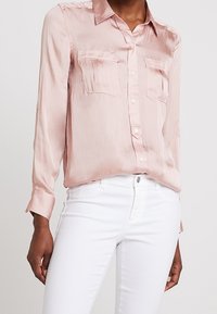 Ljusrosa satinblus med knappupptill, krage och två bröstfickor, i kombination med vita jeans. Långa ärmar med knappmanschetter.