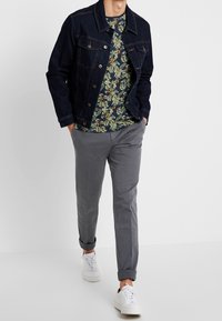 Man loopt met handen in zijn zakken, draagt een donker dennenjack, een groene bloemenhemd, grijze opgerolde broek en witte sneakers.