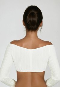 Weiße gestrickte Cropped-Bluse mit einem strukturierten Rautenmuster, schulterfreiem Design und langen Ärmeln. Die Rückansicht zeigt den Hautton und eine Haarebun.