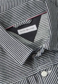 Gros plan sur le col d'une chemise rayée noire et blanche avec une étiquette Tommy Hilfiger et un bouton affichant le nom de la marque.