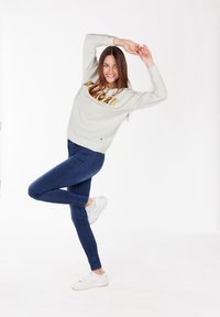 Sudadera gris claro con letras doradas, combinada con jeans de mezclilla oscuros y zapatillas blancas. El modelo tiene el cabello largo y posa de manera dinámica.