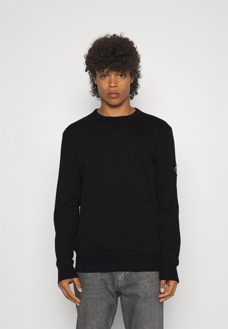Jong persoon met krullend haar, gekleed in een simpele zwarte longsleeve en grijze spijkerbroek, staand tegen een witte achtergrond.